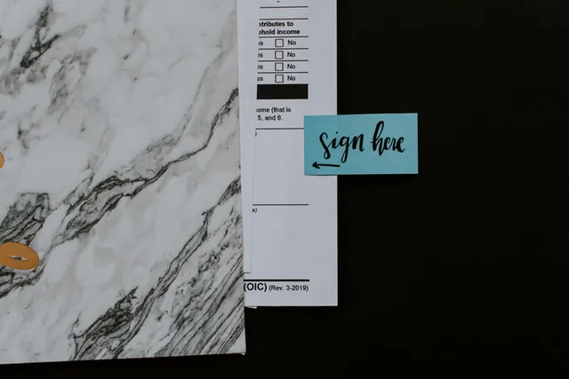 A sticky note with a sign here written on it near blank space on a printed contract.