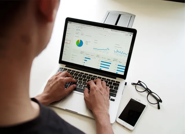 A person calculating their finances on a laptop that's located on a desk next to a pair of glasses and a smartphone.